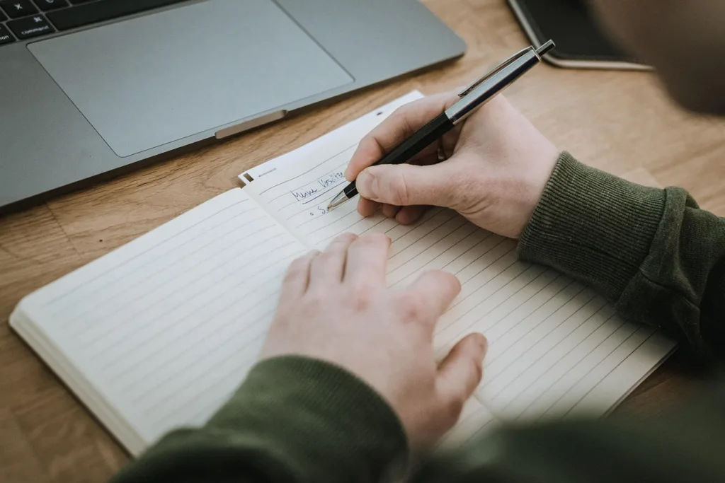 Dłonie osoby zapisującej notatki w zeszycie obok laptopa na drewnianym biurku.